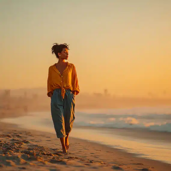 A young woman with long, natural hair walking barefoot on a California beach during golden hour.